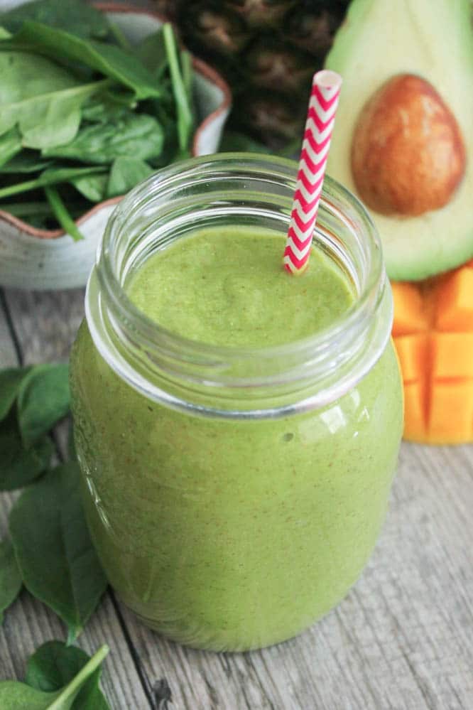 Mason jar filled with tropical green smoothie with a red and white straw. The jar is surrounded by a halved avocado, mango, and a bowl of spinach.