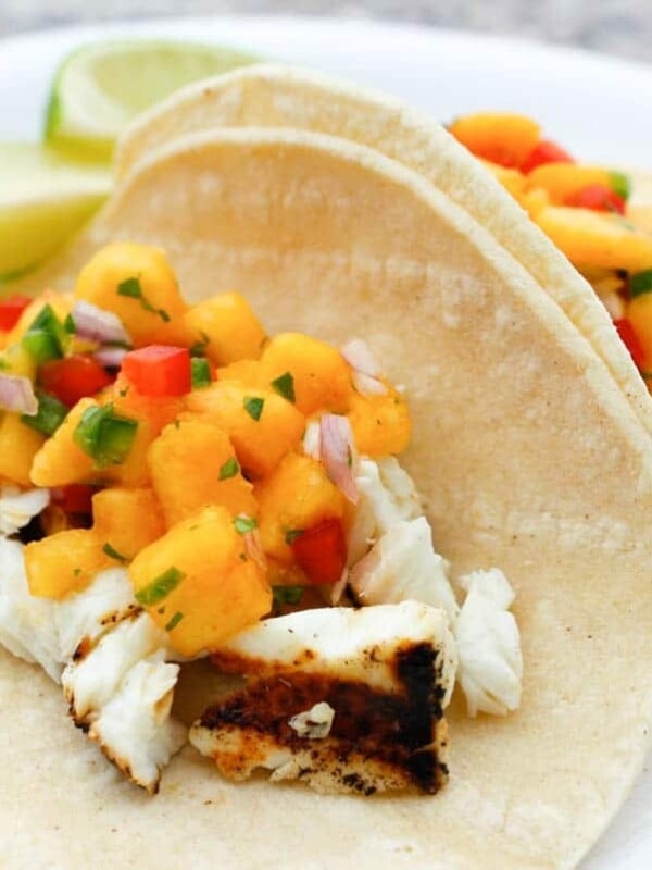 Close up of tacos filled with grilled halibut filet and peach salsa with lime wedges on the side.