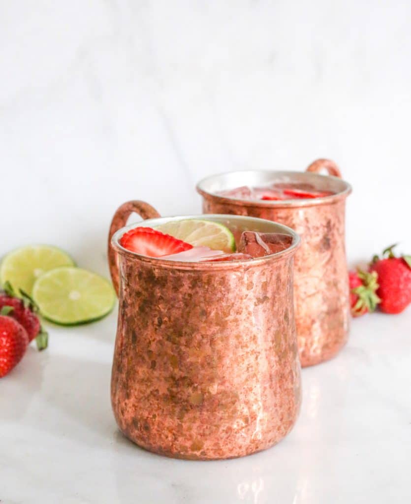Two Strawberry Mexican Mules in Moscow Mule mugs surrounded by strawberries and lime slices.
