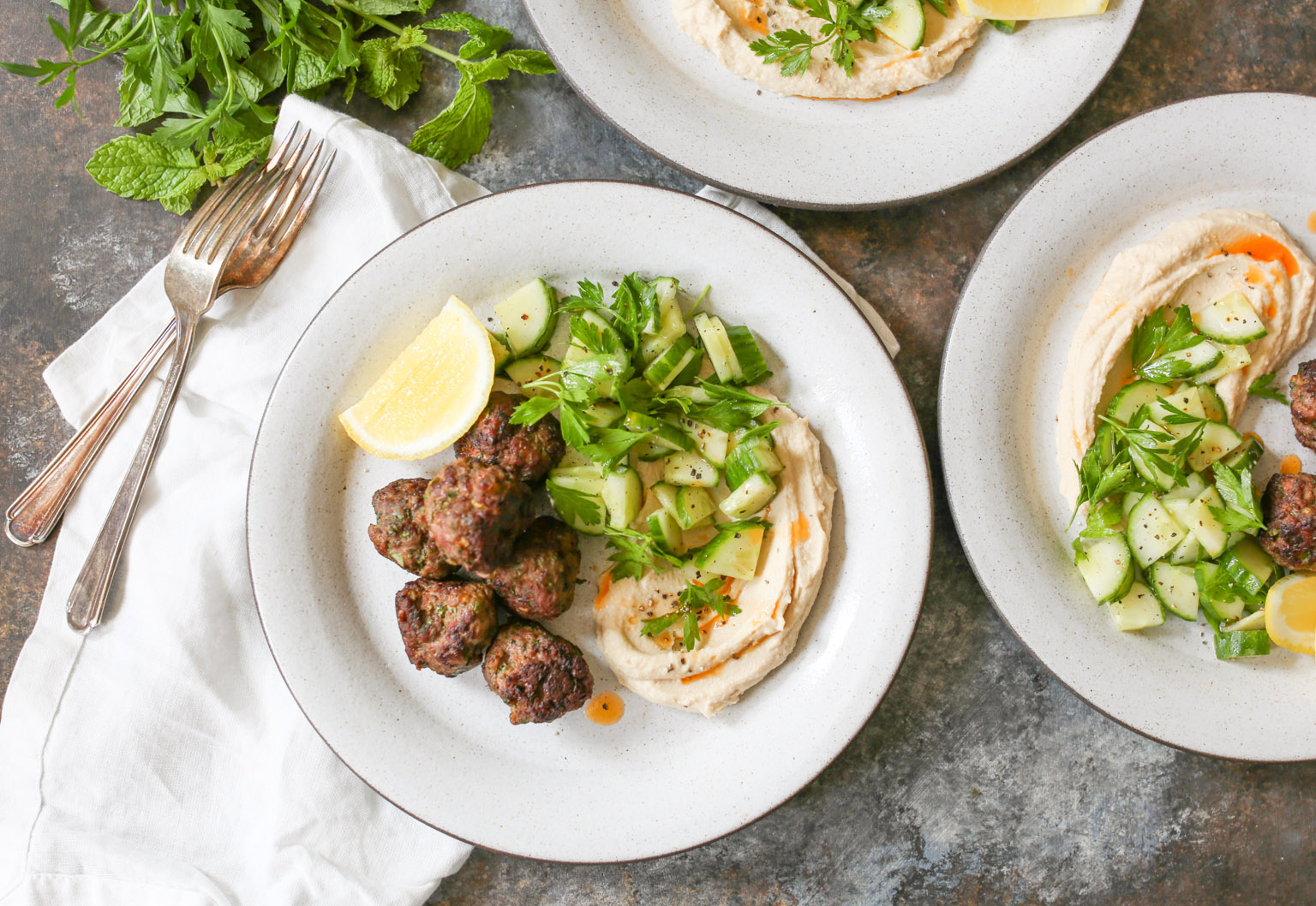 Overhead view of plated lamb kofta with hummus, cucumber salad, and lemon with napkins and forks.