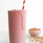 Light red PB&J smoothie in a tall glass with a red and white spiral straw and a bowl of peanut butter in the background.