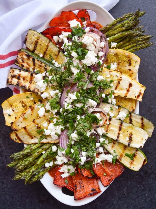 Overhead view of a platter of grilled zucchini, yellow squash, bell pepper, asparagus, and red onion topped with crumbled feta cheese and chopped herbs.
